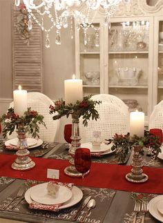 the table is set for christmas dinner with red and silver place settings, white china dishes, candles and greenery