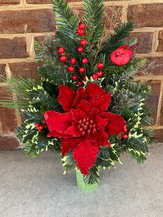 a green vase filled with red flowers and greenery on top of a sidewalk next to a brick wall