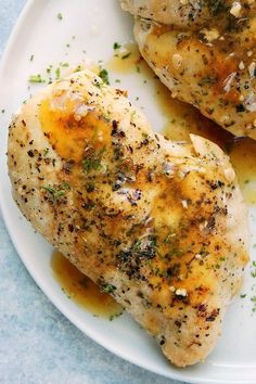 two pieces of chicken on a white plate covered in herbs and seasoning next to sauce