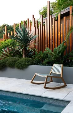 a wooden rocking chair sitting next to a swimming pool surrounded by greenery and trees