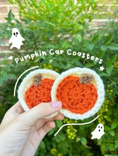 a hand holding two crocheted pumpkin coasters in front of some bushes and trees