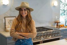 a woman wearing a hat standing in front of a stove top oven with her arms crossed