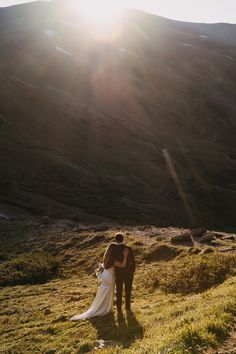 a bride and groom standing in the sun on top of a hill with mountains behind them