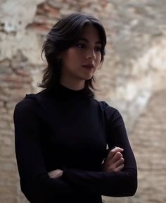 a woman standing in front of a brick wall with her arms crossed and looking off to the side