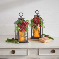 two lanterns with red berries and greenery are sitting on a table next to a present