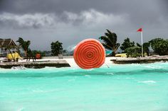 a large red object floating in the ocean next to a sandy beach with palm trees