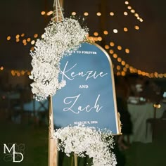 a wedding sign with baby's breath hanging from the side and lights in the background