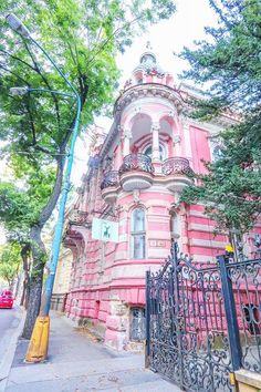 an old pink building on the corner of a city street with iron fence and trees