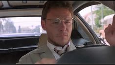 a man wearing glasses sitting in the driver's seat of a car while talking to someone