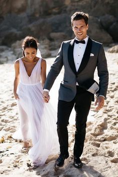 a bride and groom walking on the beach holding hands, dressed in tuxedo