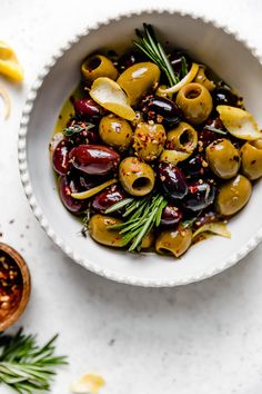 a bowl filled with olives and herbs on top of a white table next to some lemon wedges