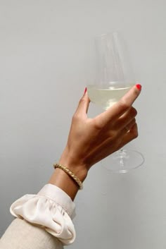 a woman's hand holding a wine glass with white wine in it, against a gray wall