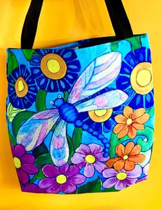 a blue bag with flowers and a dragonfly painted on the front is hanging against a yellow wall