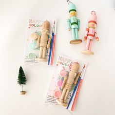 three wooden toy figures are on the table next to markers and pencils, with a small christmas tree in the background