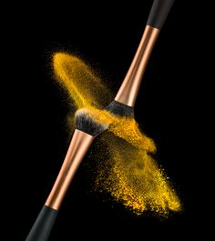 two brushes with yellow powder flying out of them on a black background, one brush has gold flecks in it