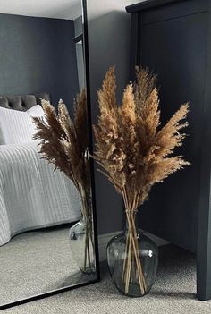 dried flowers in a vase on the floor next to a mirror with a bed in the background