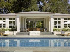 an outdoor pool with chairs and umbrellas next to the side of a house that is surrounded by trees