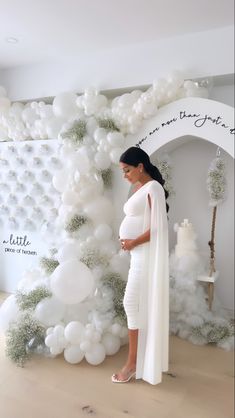 a pregnant woman standing in front of a backdrop with white balloons and greenery on it