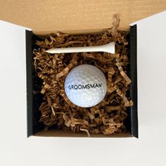 a golf ball in a cardboard box with wood shavings around it and the word groomsman on top