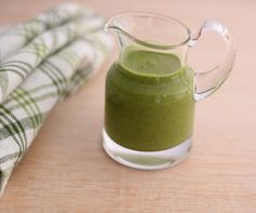 a glass pitcher filled with green smoothie on top of a wooden table next to a towel