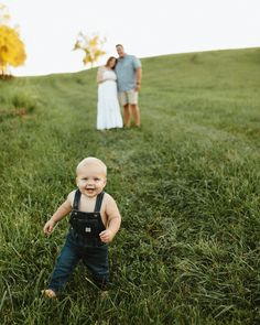 a baby boy standing in the grass with his mom and dad behind him on a hill