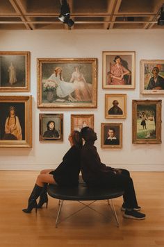 two people are sitting on a bench in front of some paintings and pictures hanging on the wall