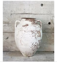 an old white vase sitting on top of a wooden table next to a wall with holes in it