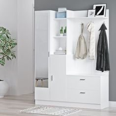 a white cabinet with some coats hanging on it's doors and shelves next to a potted plant