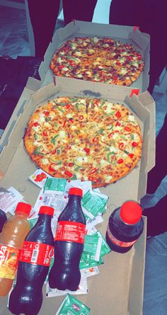 two pizzas in boxes with drinks on the table next to each other and bottles of soda