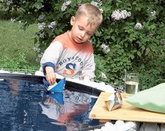 a young boy is playing with paper boats in an outdoor swimming pool that's surrounded by flowers and shrubs