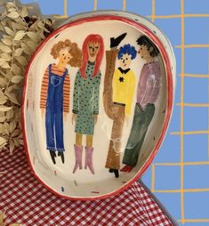 a plate with people painted on it sitting on a checkered tablecloth next to a pine cone