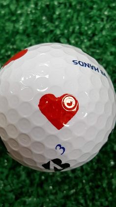 a white and red golf ball sitting on top of a green carpeted field with an i love heart sticker on it's side