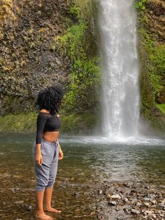 a woman standing in front of a waterfall with her back to the camera and looking at it