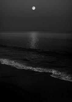 the moon is shining over the ocean on a black and white night with waves coming in