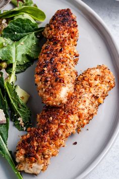 two pieces of chicken on a white plate next to a green salad and some fork