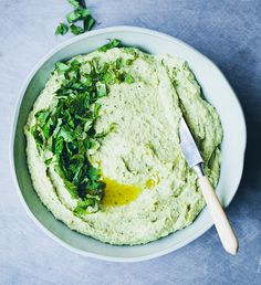 a white bowl filled with green hummus and greens next to a knife on a blue surface