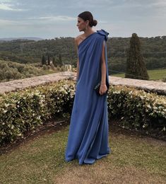 a woman in a blue dress standing on top of a lush green field next to bushes