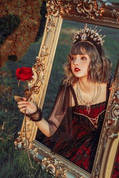 a woman in a red dress holding a rose and looking into a gold framed mirror