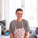 a woman standing in front of a kitchen sink with an apron over her shoulder and smiling at the camera