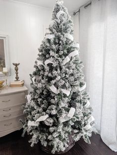 a white christmas tree in a basket on a wooden floor next to a window with curtains