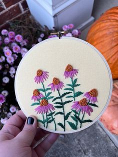 a person holding up a small embroidery project with purple flowers on it and orange pumpkins in the background