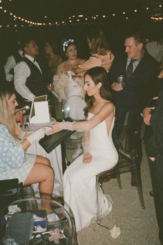 a woman in a white dress is sitting at a table with other people around her