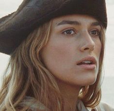 a woman with long hair wearing a black hat and looking off to the side while standing in front of an ocean