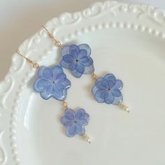 three blue flowers are on a white plate with pearls and gold ear wires in the shape of four petals