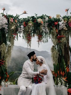 a bride and groom sitting on a bench in front of an arch with floral decorations