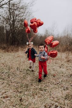 two young children holding red balloons and walking through a field with trees in the background