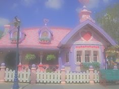 a pink and purple house with a heart shaped window on the roof, next to a white picket fence