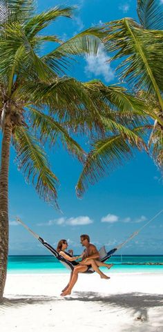 a man and woman sitting in a hammock under a palm tree on the beach