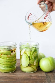 a person pouring dressing into jars filled with green vegetables and apples on a wooden cutting board