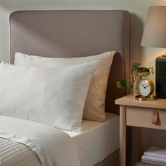 a bed with white sheets and pillows next to a night stand, lamp and nightstand